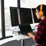 Woman programming on a laptop at a standing desk in an office with large windows.